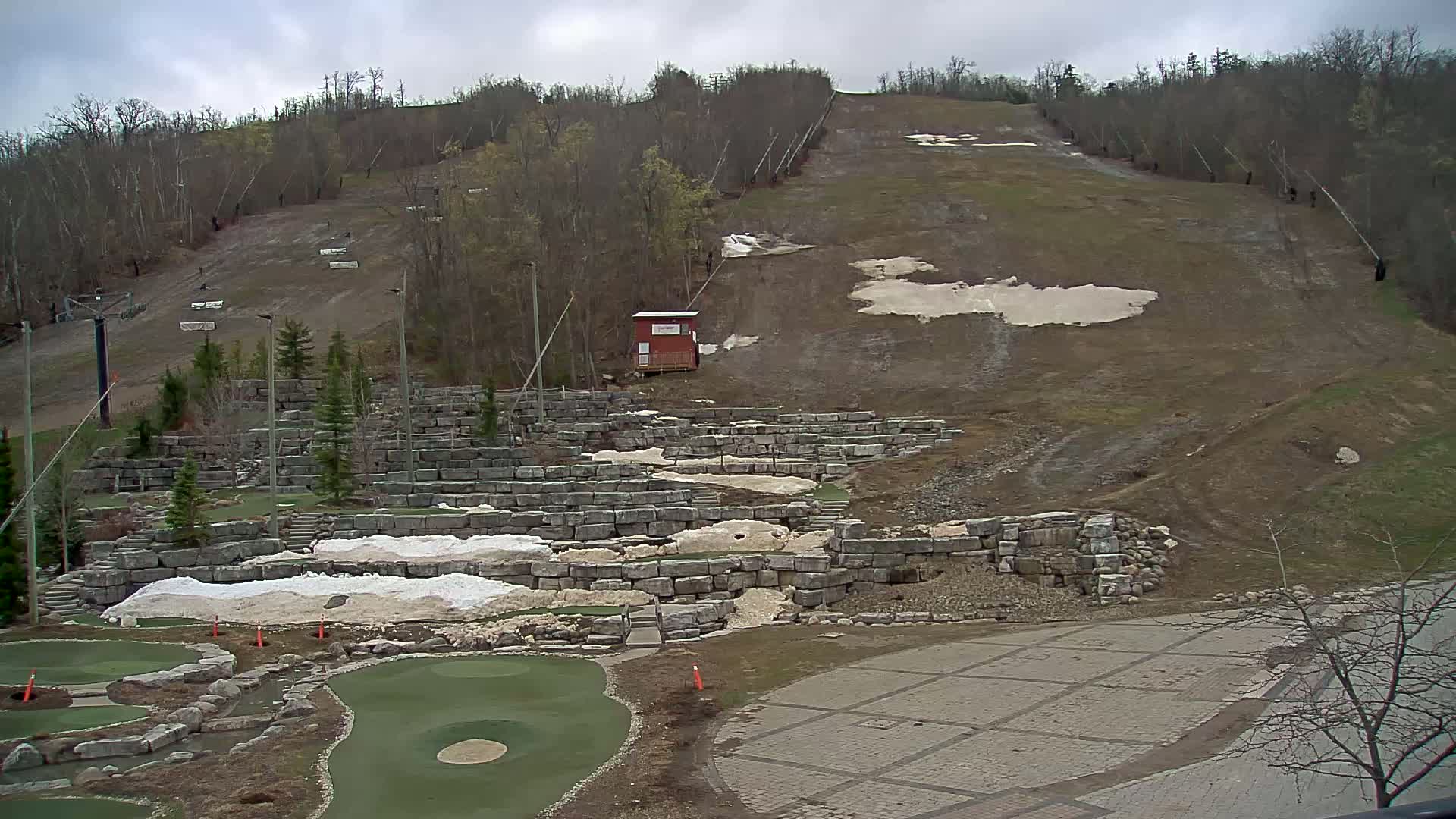 A mostly barren ski slope with patches of snow overlooks a stone-walled mini golf course on an overcast day.