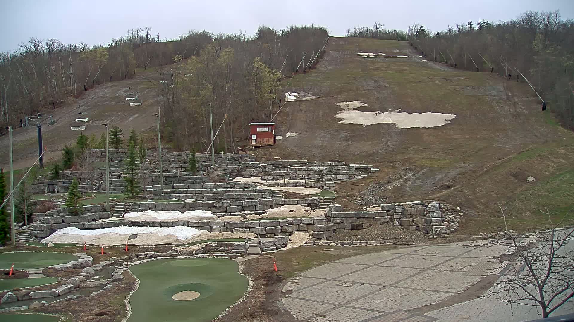 A ski slope with some remaining snow, a small building, and a stone-walled mini golf course below, all under an overcast sky.