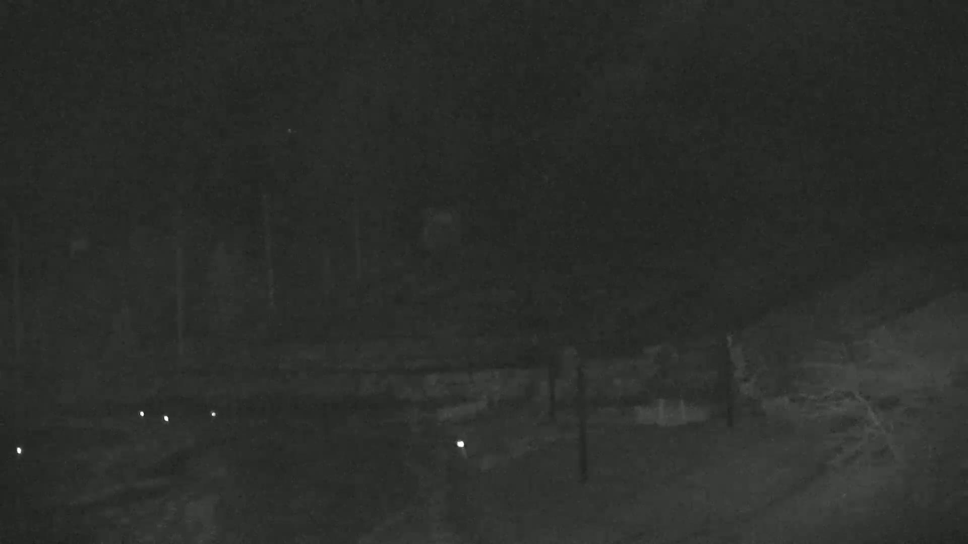 A dark, nighttime scene shows a dimly lit area with a few small lights near a low wall and some trees or bushes in the background.