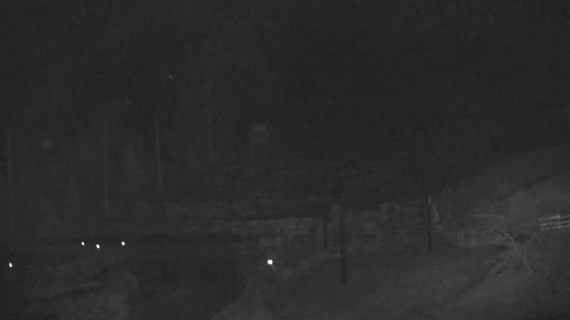 A nighttime, low-light image shows a dark, outdoor scene with dimly lit structures and a few small lights on the ground.