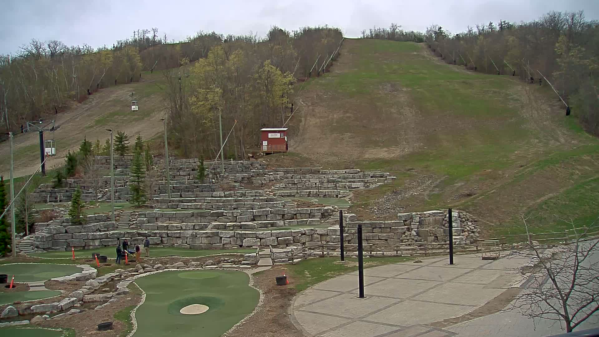 An outdoor scene on an overcast day shows a ski hill with a chairlift, a small building, and a stone retaining wall surrounding a miniature golf course.