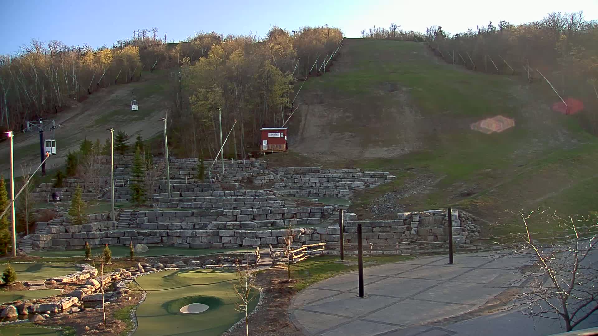 A grassy hillside with a ski lift and stone retaining walls featuring a miniature golf course, all under a clear, sunny sky.