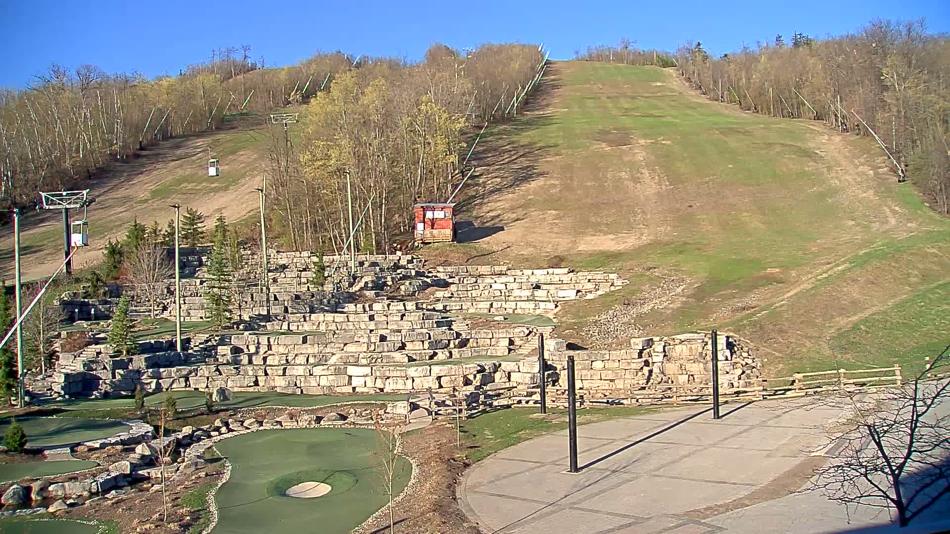 A grassy ski hill with chairlifts and a stone-walled miniature golf course below on a sunny day.