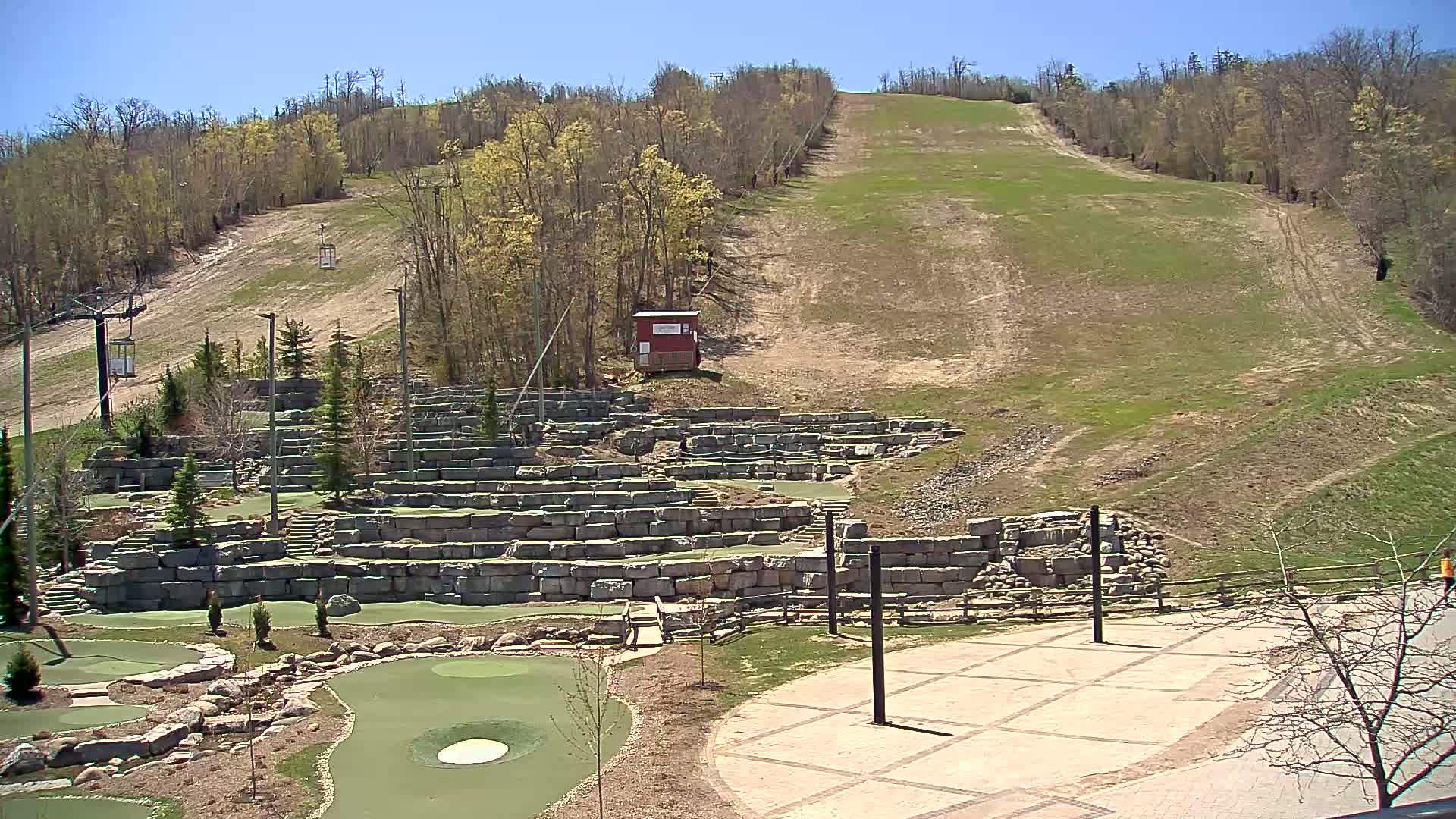 A grassy ski slope with a chairlift rises behind a stone-walled miniature golf course on a clear, sunny day.