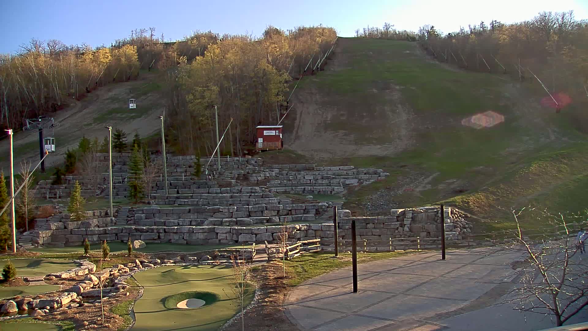 A grassy hillside with a ski lift and stone retaining walls, featuring a miniature golf course in the foreground, under a clear, sunny sky.