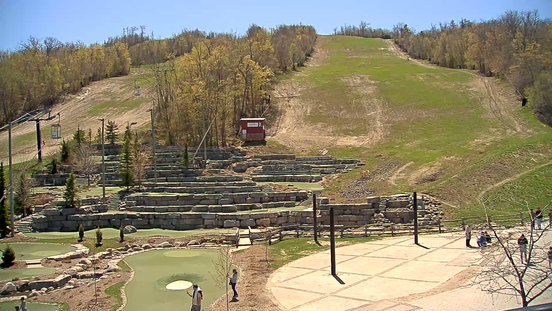 A grassy ski hill slopes upward behind a stone mini-golf course, under a clear blue sky.