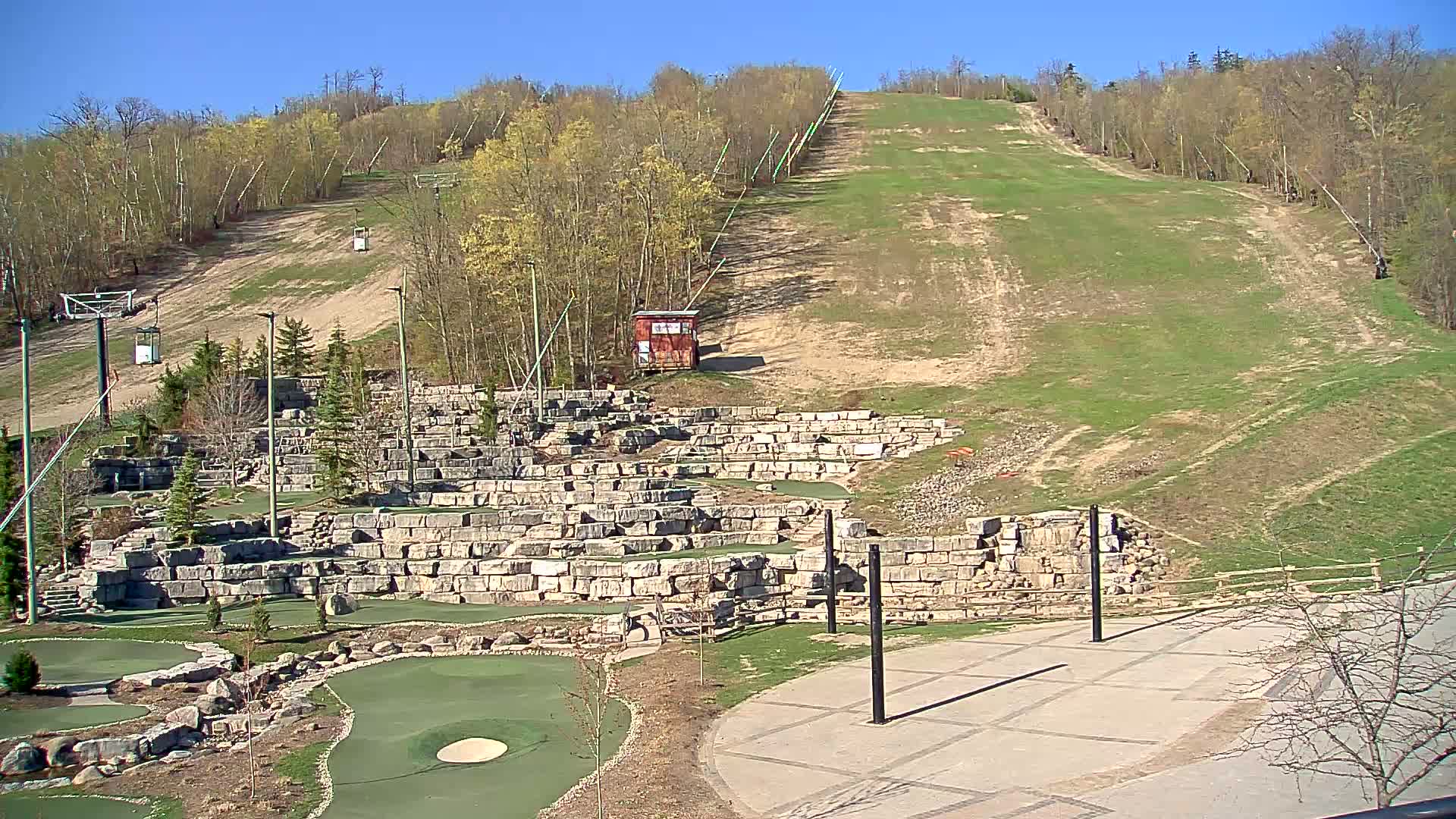 A grassy ski hill with a chairlift is visible in the background, while a stone-walled miniature golf course and paved area are in the foreground under a clear blue sky.
