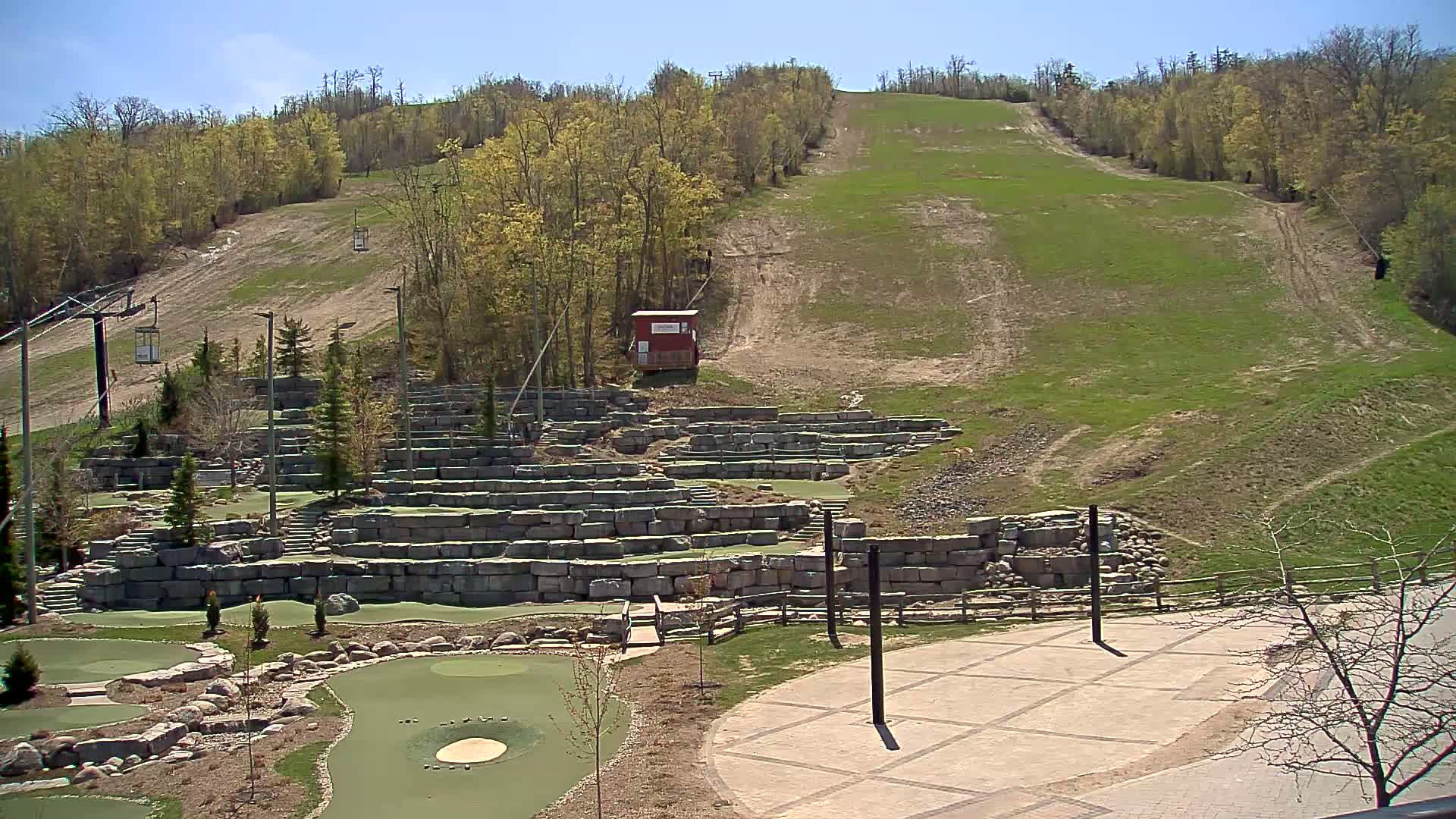 A grassy hillside with a ski lift and a stone-walled mini golf course below on a sunny day.