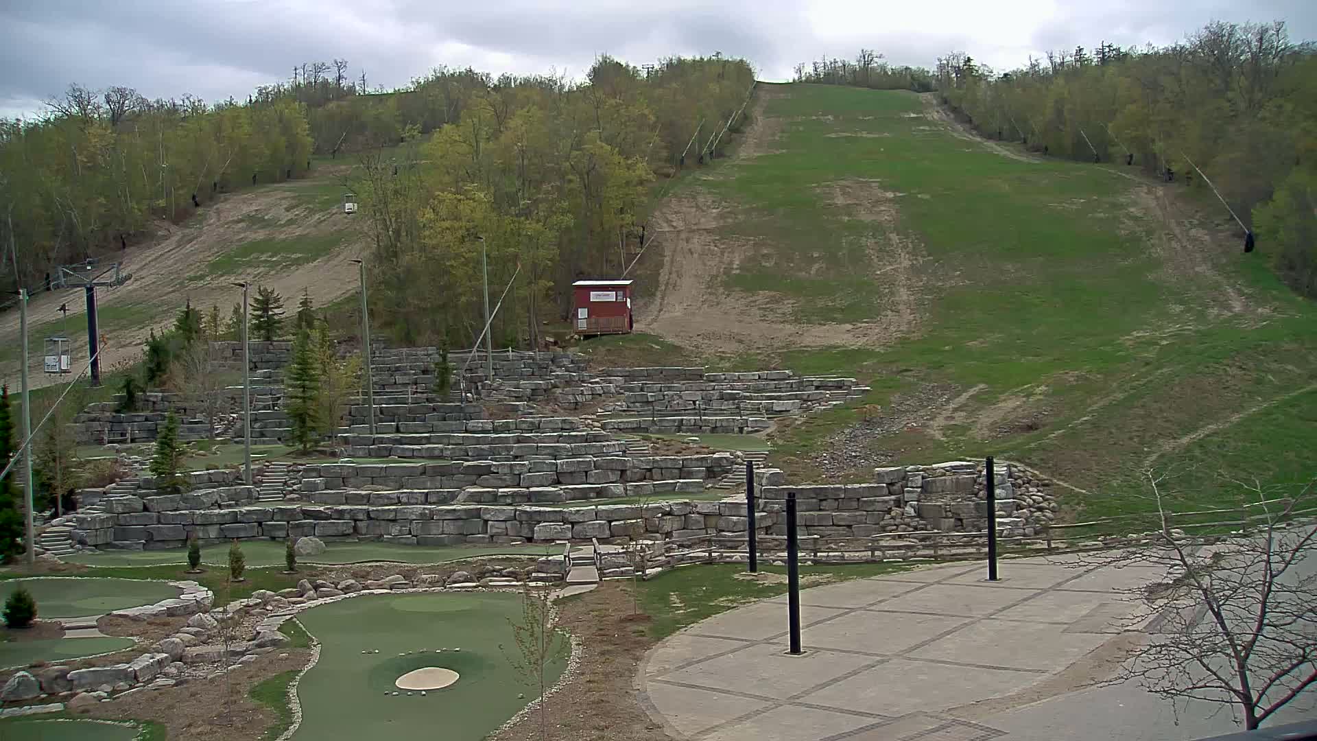 A grassy ski hill with a chairlift, a small red building, and a stone-walled miniature golf course below, under a cloudy sky.
