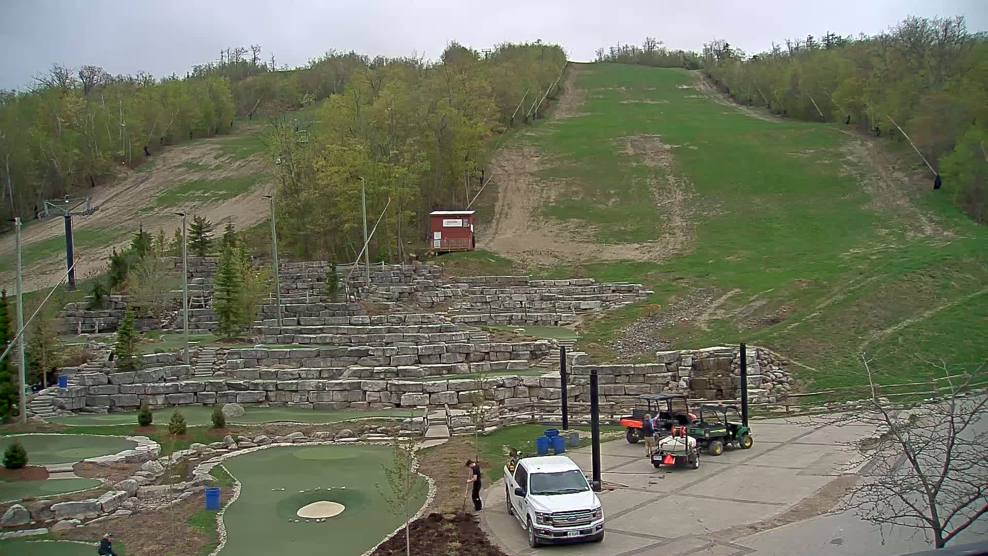 A grassy ski slope is visible behind a stone-walled miniature golf course with several utility vehicles parked in a paved lot under an overcast sky.