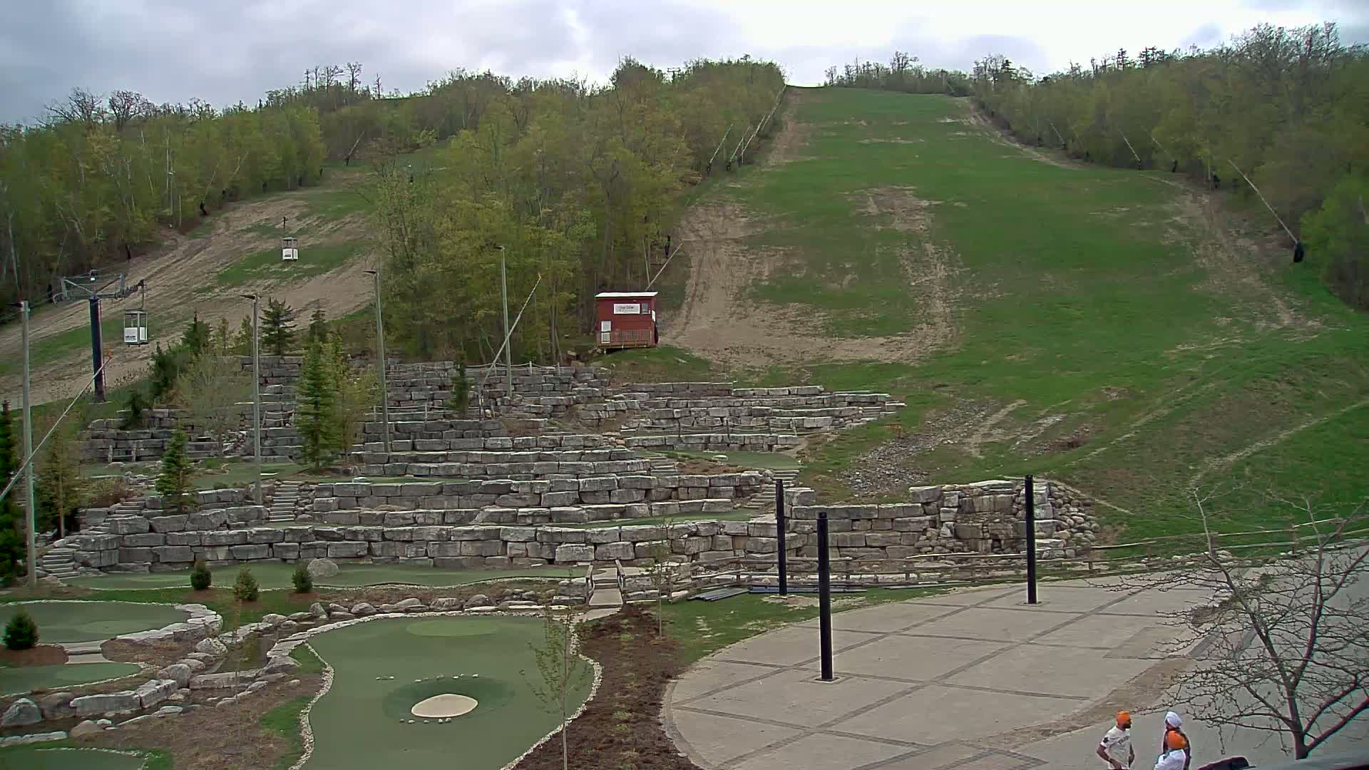 A grassy hillside with a ski lift and stone retaining walls forms a backdrop for a paved area and a miniature golf course, all under a cloudy sky.