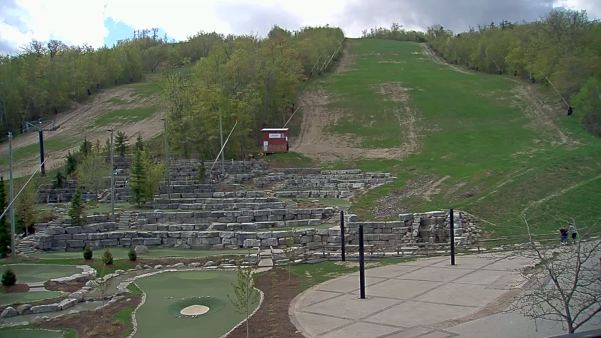 A grassy hillside with a ski lift, a small building, and a stone-walled mini-golf course below, under a cloudy sky.
