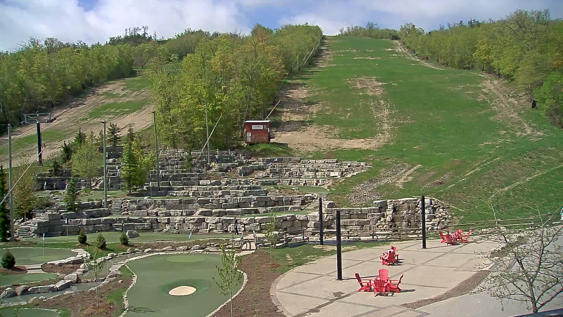 An outdoor scene on a partly sunny day shows a grassy ski hill in the background, with a mini-golf course and patio furniture in the foreground.