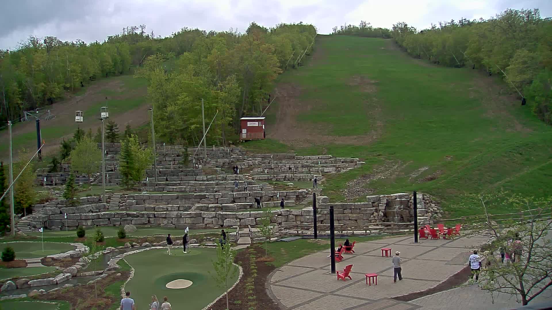 An outdoor mini-golf course with a grassy ski hill in the background under overcast skies.