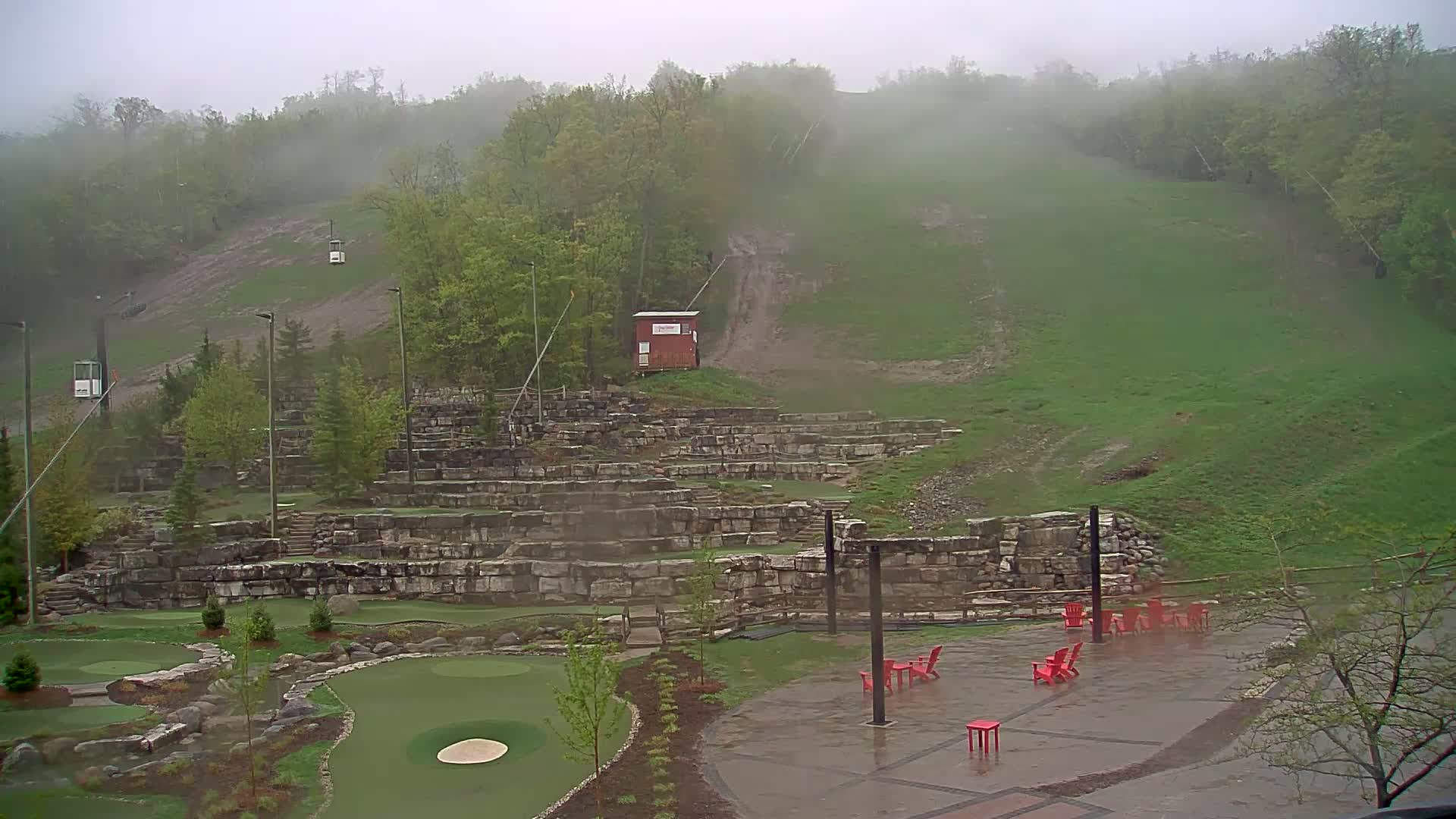 A mini golf course with stone retaining walls is situated below a grassy hill with a chairlift and a small building, all under a foggy, rainy sky.