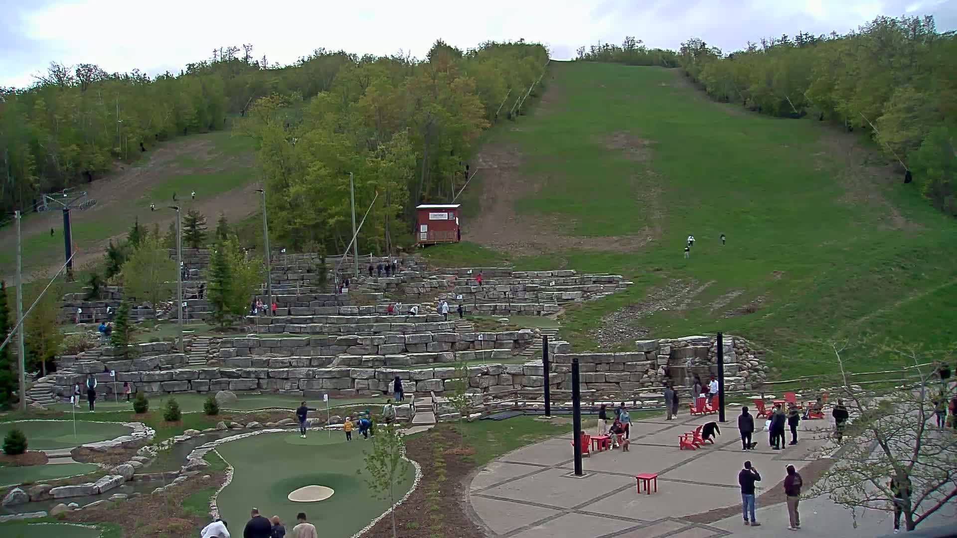 An outdoor mini-golf course with stone walls and green artificial turf is situated at the base of a grassy hill covered in trees under a cloudy sky.