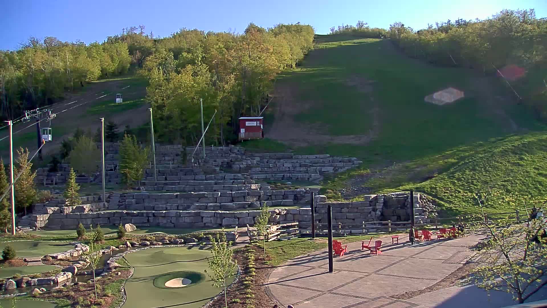 A grassy hillside with a ski lift, stone retaining walls forming a tiered landscape with a miniature golf course in the foreground, and red chairs on a patio under a clear blue sky.