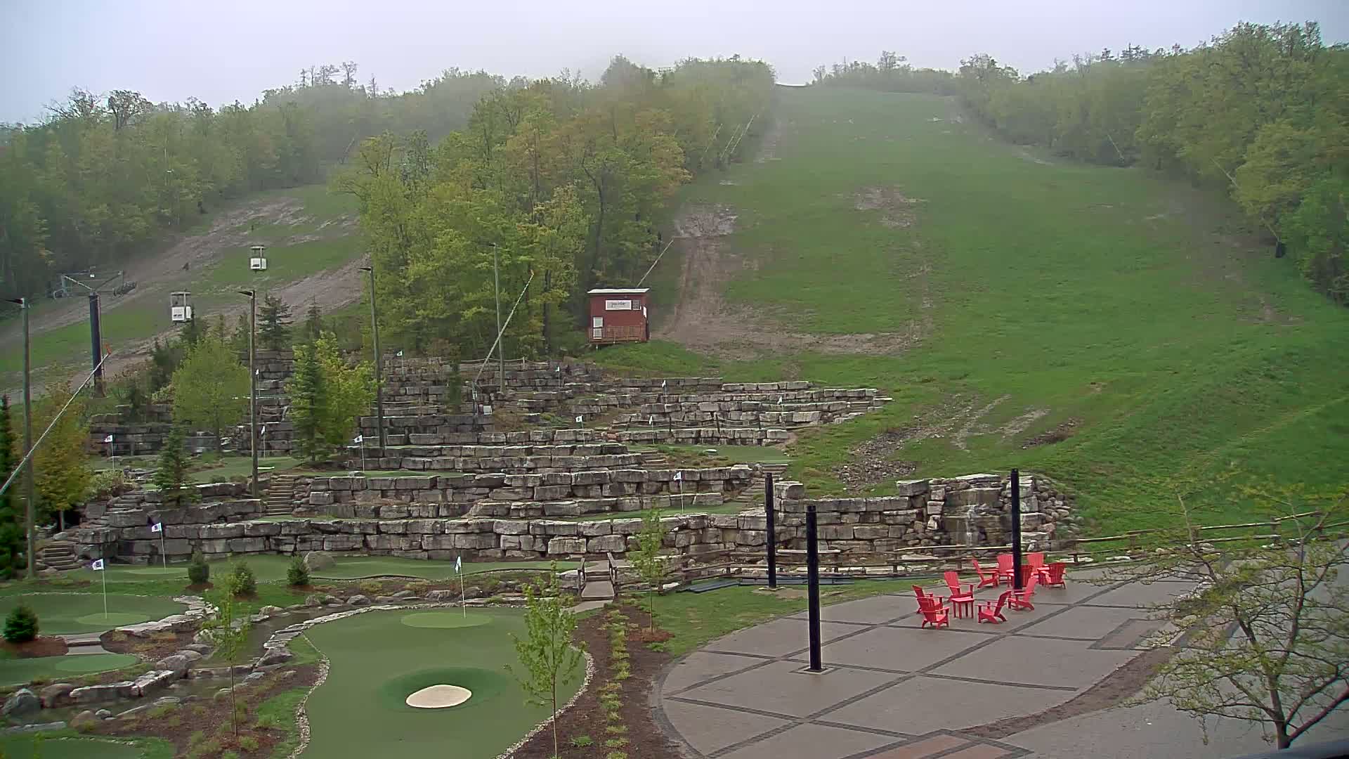 A hillside covered in green grass and trees, featuring a stone-walled mini golf course and a patio with red chairs, is viewed on an overcast day.