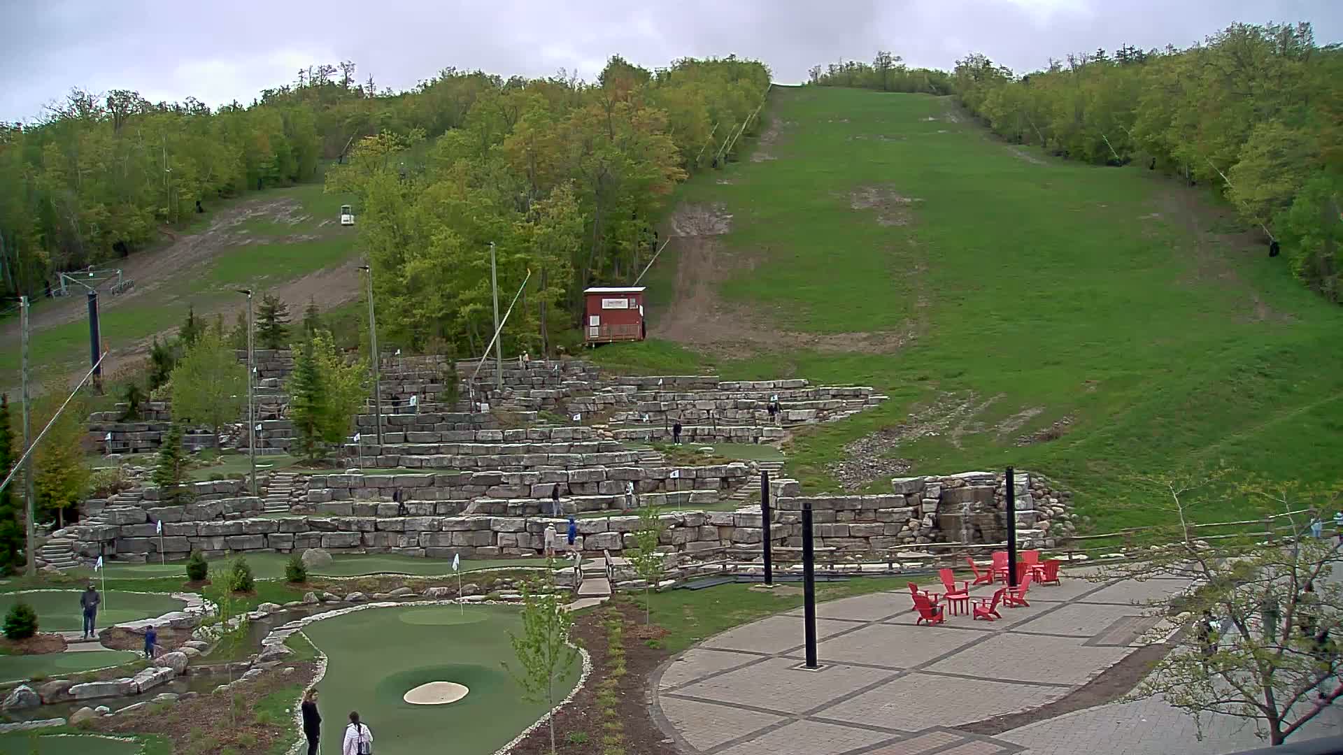 An outdoor mini-golf course with stone retaining walls and a grassy hillside in the background under an overcast sky.