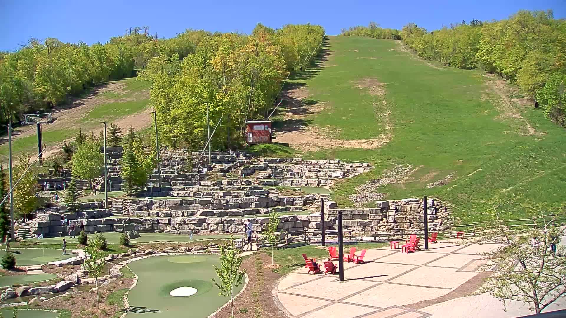 A grassy hillside with a ski lift rises behind a stone-walled miniature golf course and patio area with red chairs, all under a clear blue sky.