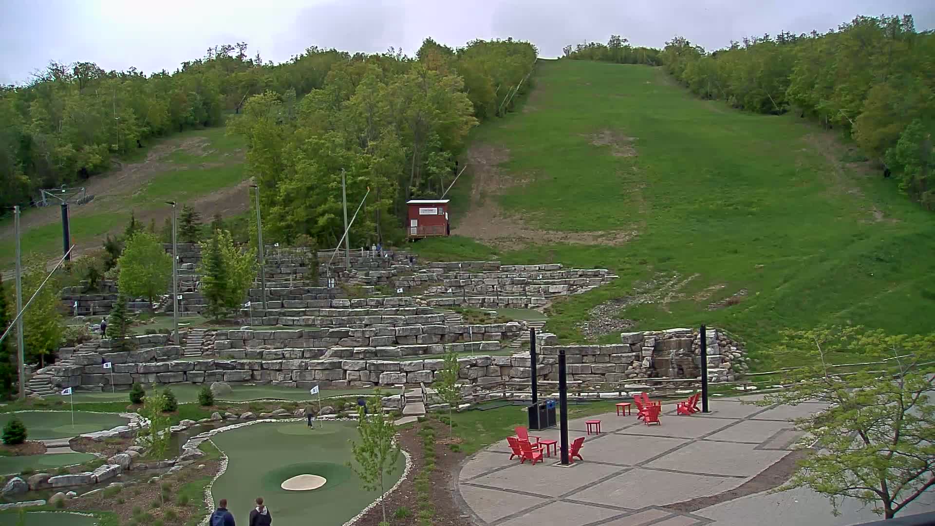 An outdoor mini golf course built into a hillside, with a grassy ski slope visible in the background under an overcast sky.