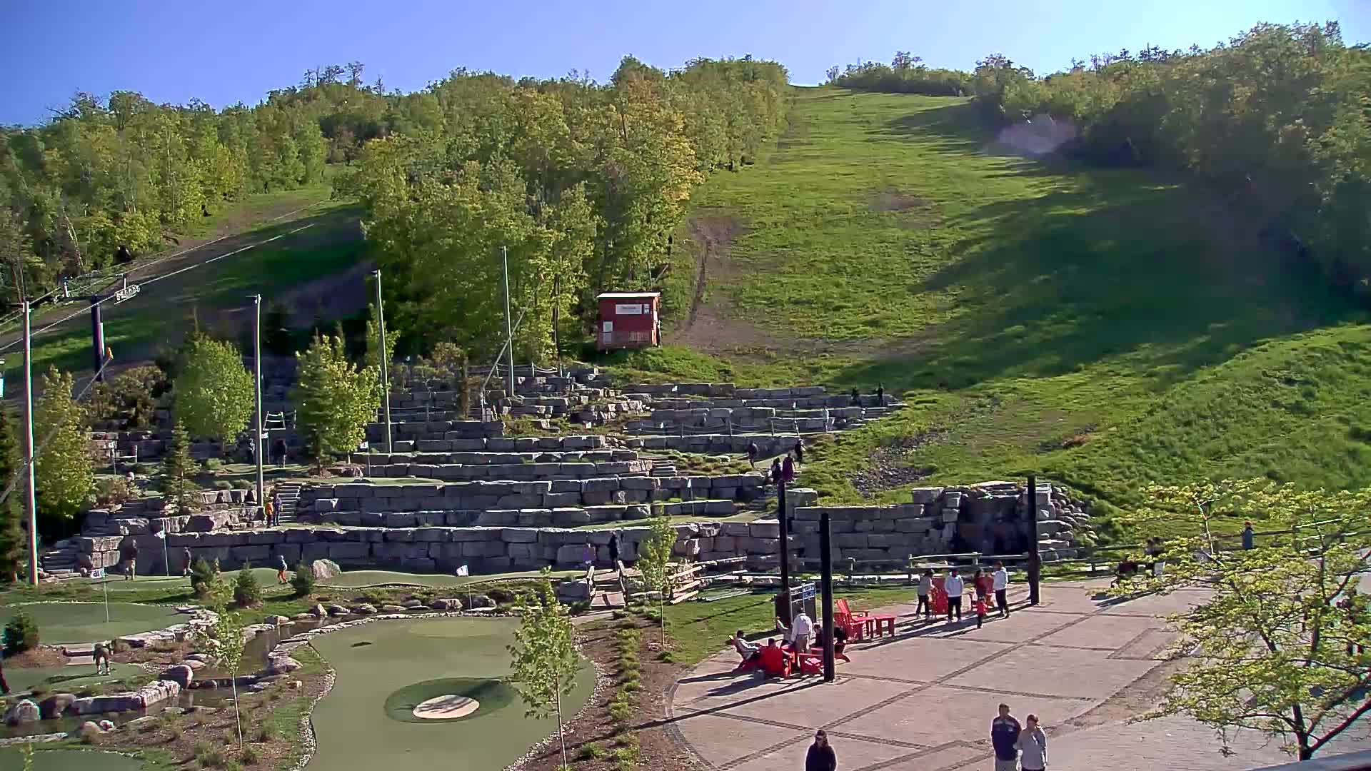 A grassy hillside with a stone mini-golf course and people relaxing in red chairs under a clear, sunny sky.
