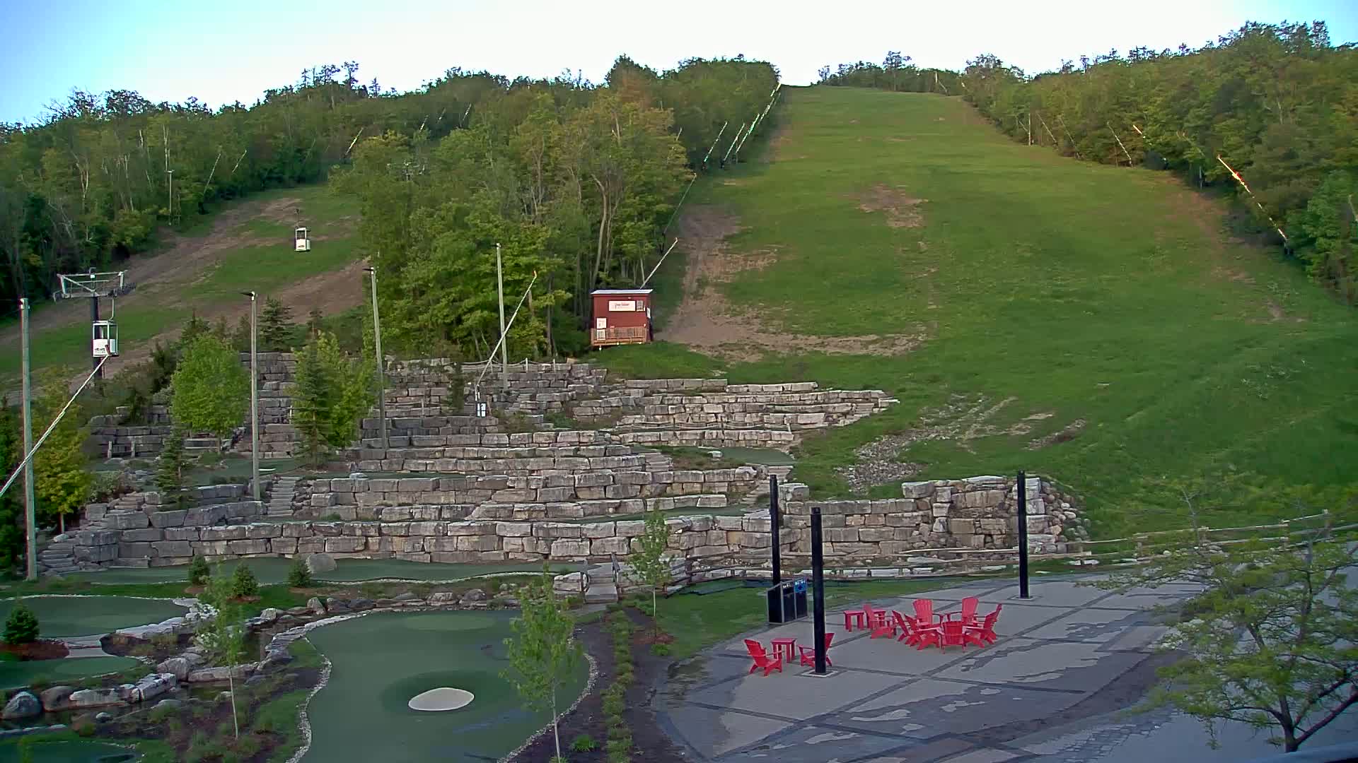 A grassy hillside with a ski lift and a stone amphitheater-like structure, featuring a miniature golf course and several red chairs in the foreground, under a clear sky.