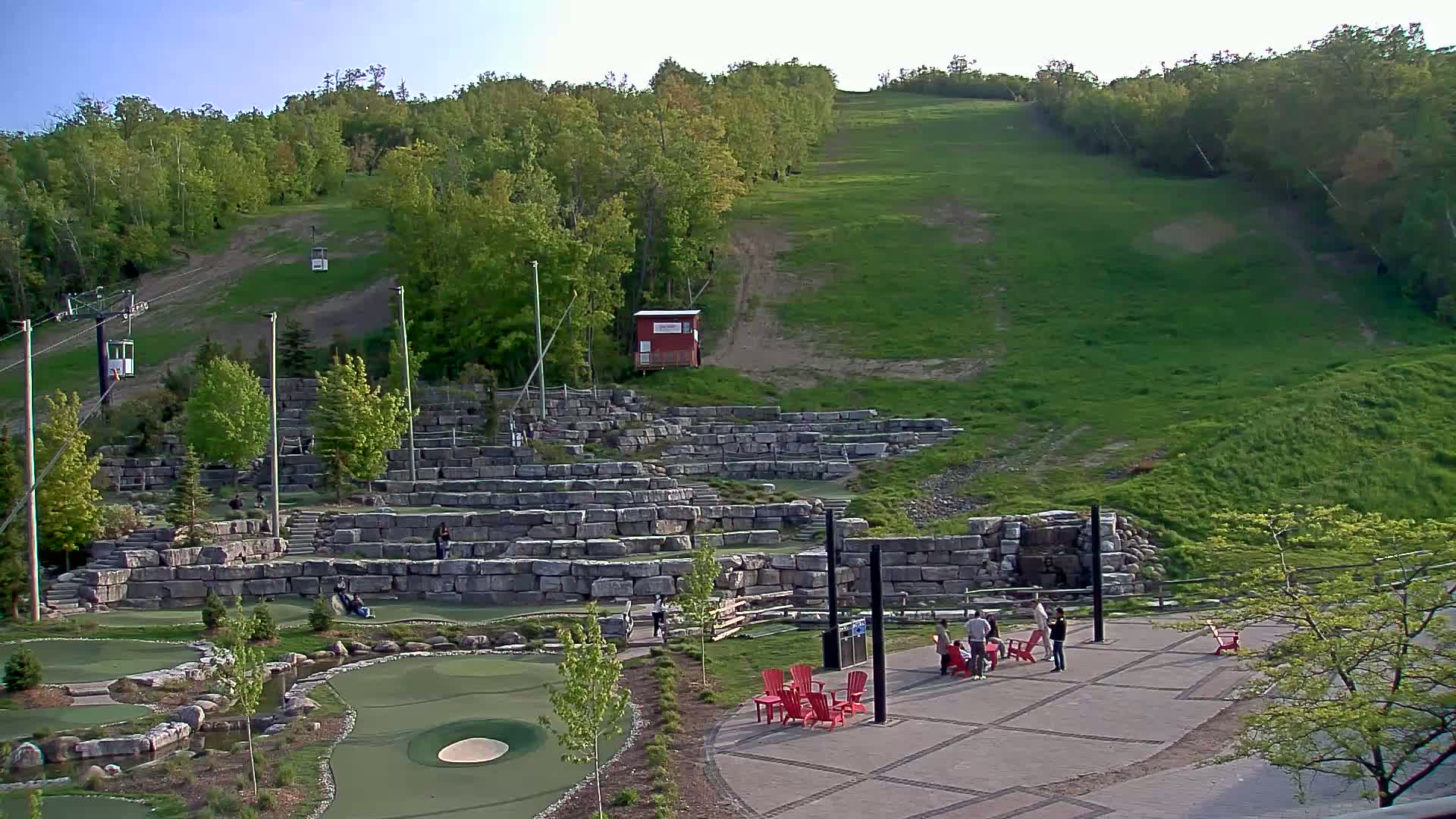A grassy hillside with a chairlift and stone structures featuring a mini-golf course and seating area under a clear sky.
