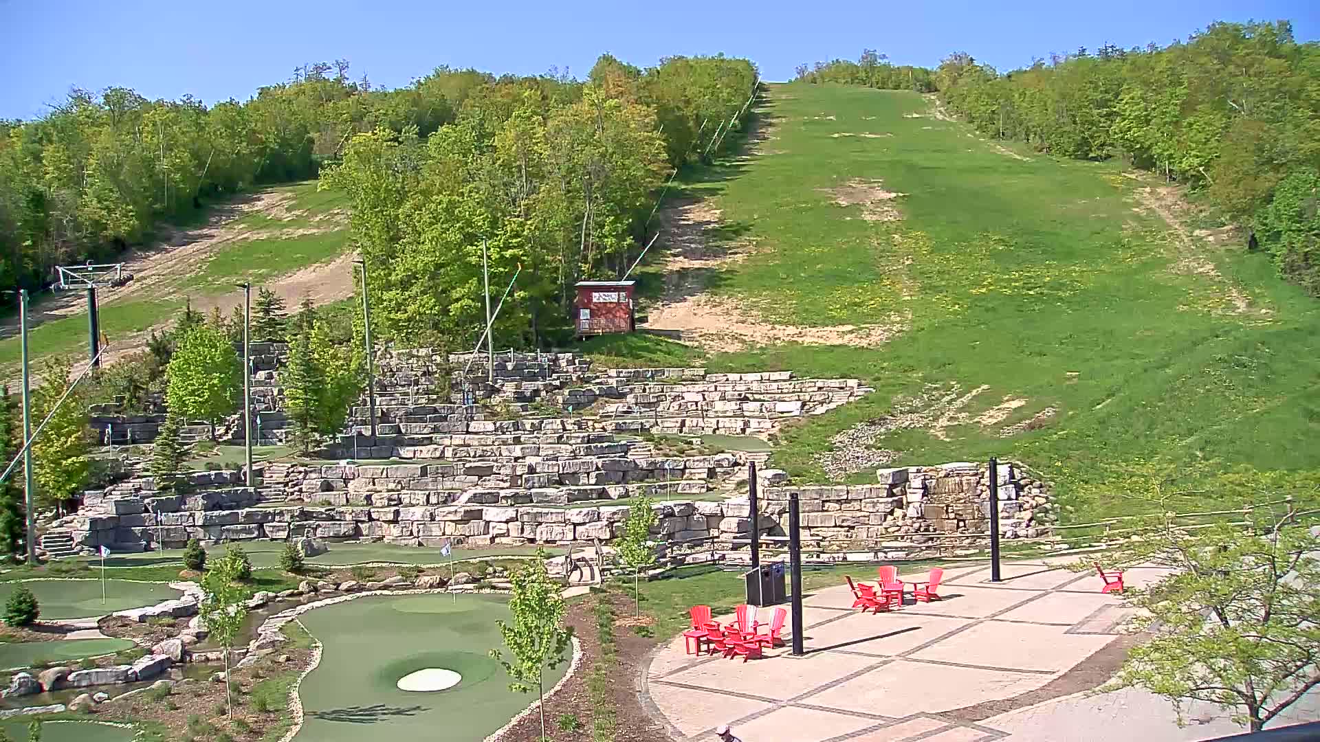 A grassy hillside slopes down to a stone-walled mini golf course and patio area with red chairs, under a clear blue sky.