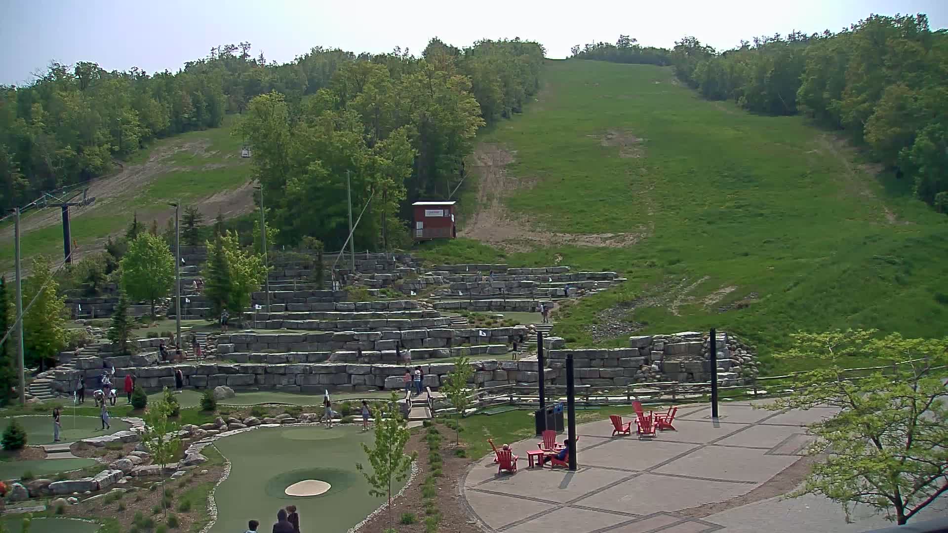 A grassy hillside with a ski lift and a mini-golf course featuring stone walls and people playing, under a clear sky.