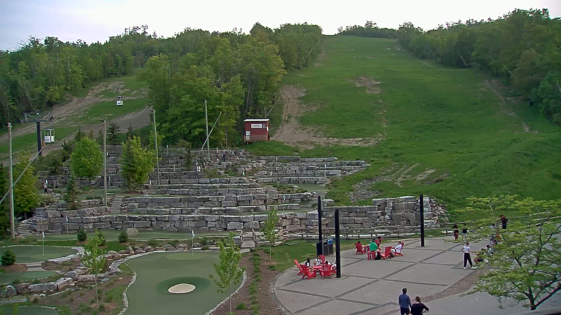 An outdoor scene under a partly cloudy sky shows a grassy hill with a ski lift and a stone-walled area with a mini golf course and people sitting at red chairs around tables.