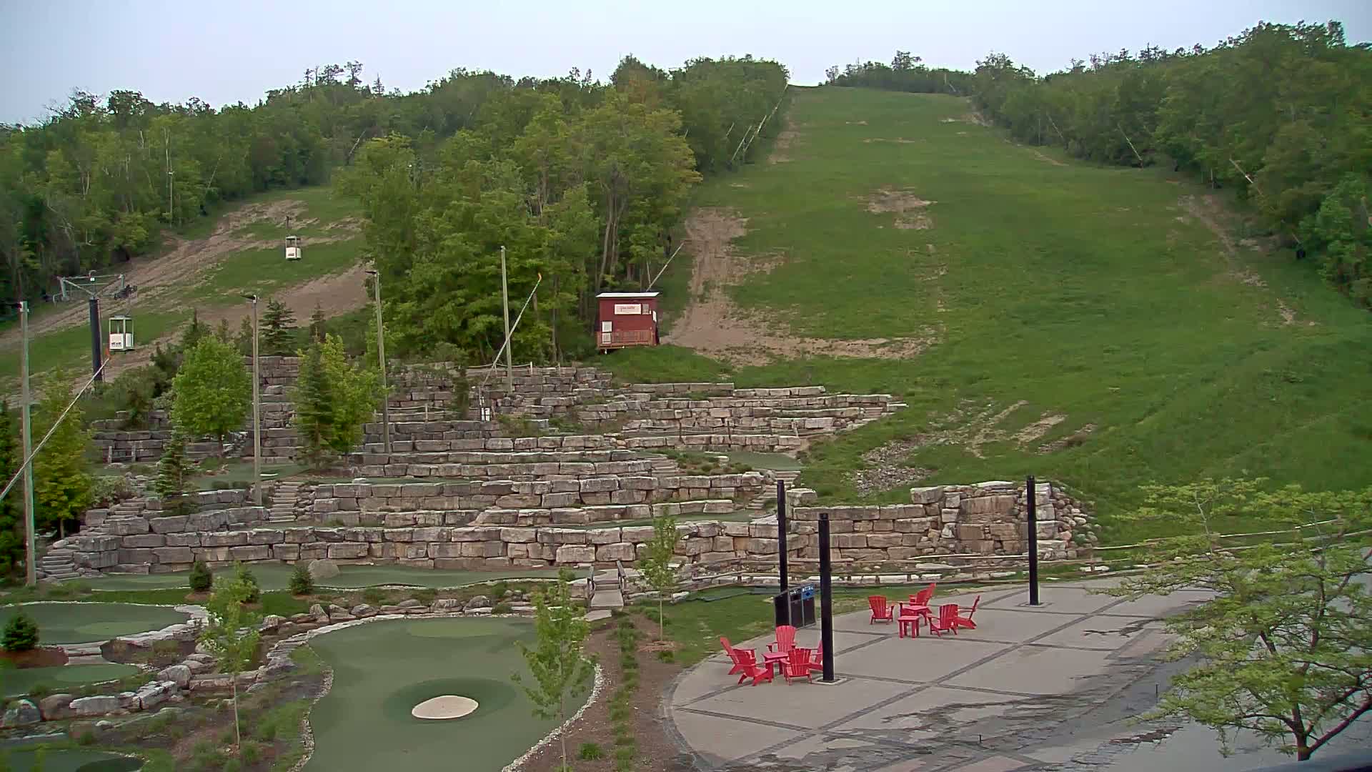 A grassy hillside with a ski lift, stone retaining walls forming tiered landscaping, and red chairs on a paved area are visible under an overcast sky.