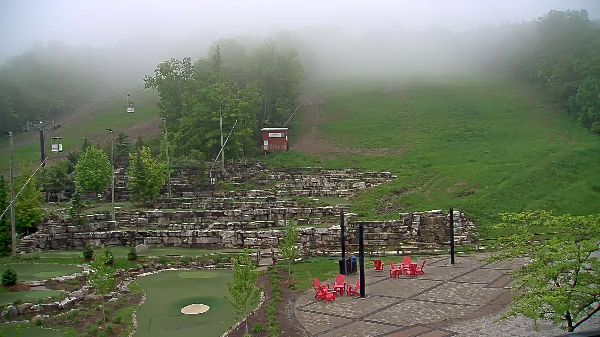A fog-shrouded outdoor mini golf course with a chairlift ascending a grassy hill in the background.