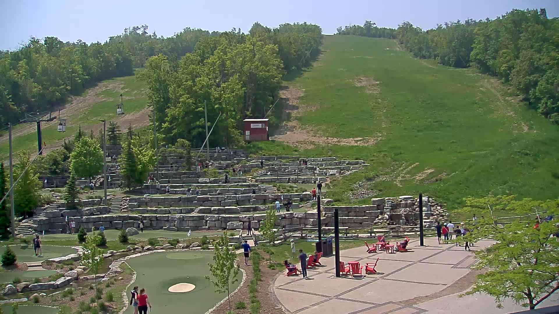 A grassy hillside with a chairlift rises above an outdoor mini-golf course with people playing on a sunny day.