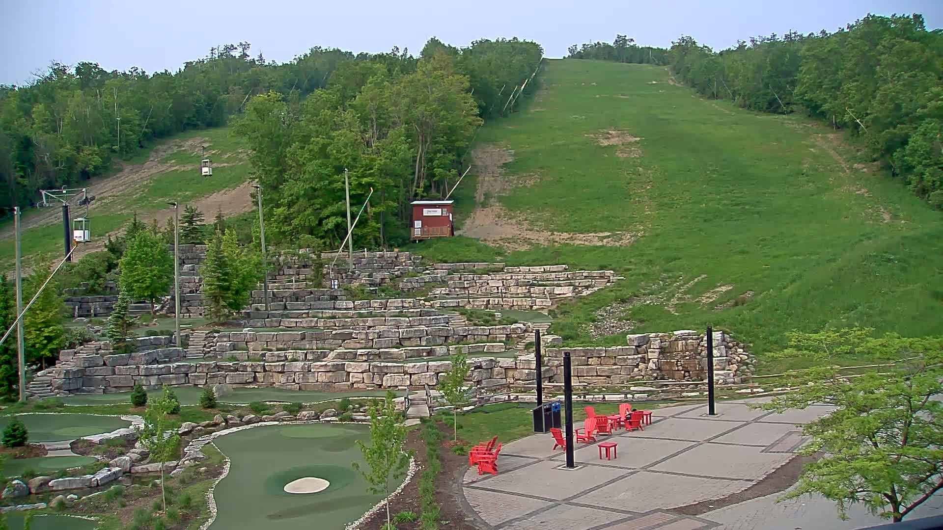 A grassy hillside with a ski lift and a stone terraced area containing a miniature golf course and red chairs under a mostly clear sky.