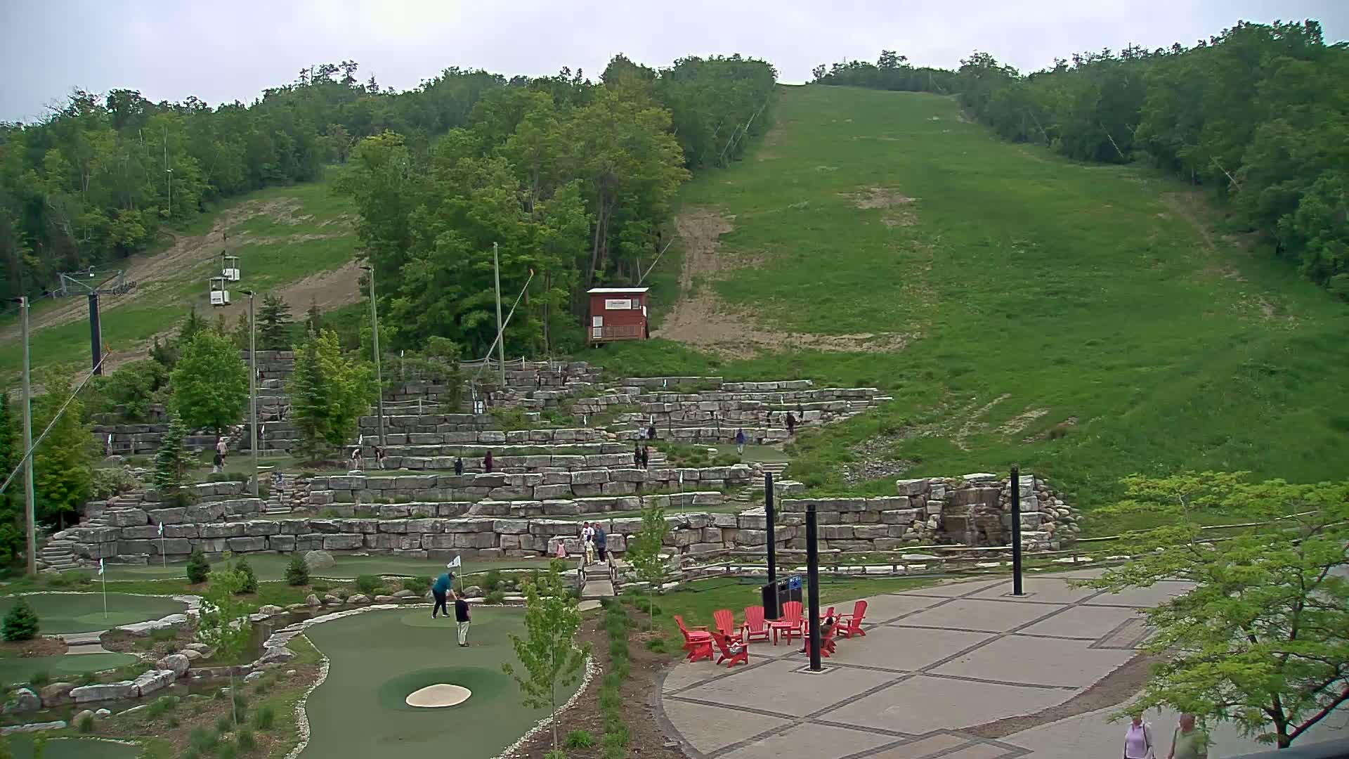 An outdoor mini-golf course with stone walls and a grassy hillside in the background under an overcast sky.