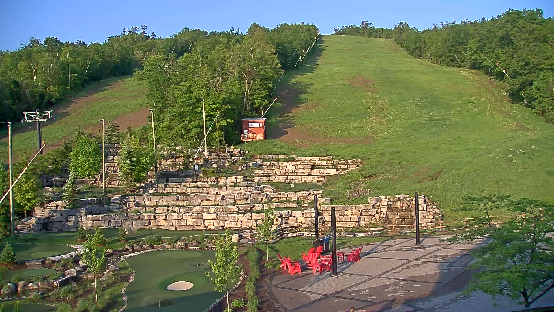 A grassy ski slope overlooks a stone amphitheater, a mini golf course, and a paved area with red chairs, all under a clear blue sky.