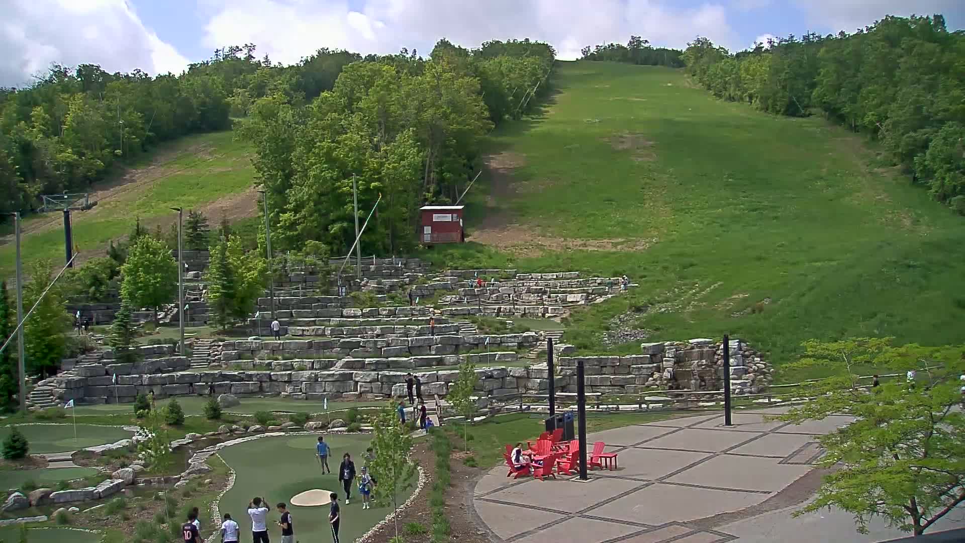 A grassy hillside, dotted with trees, overlooks a stone-walled miniature golf course with people scattered around, under a partly cloudy sky.