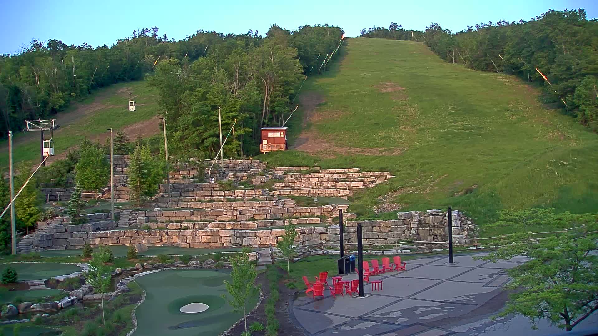A grassy hill with a ski lift and a stone amphitheater-like structure, featuring a mini golf course and red chairs, is visible under a clear sky.
