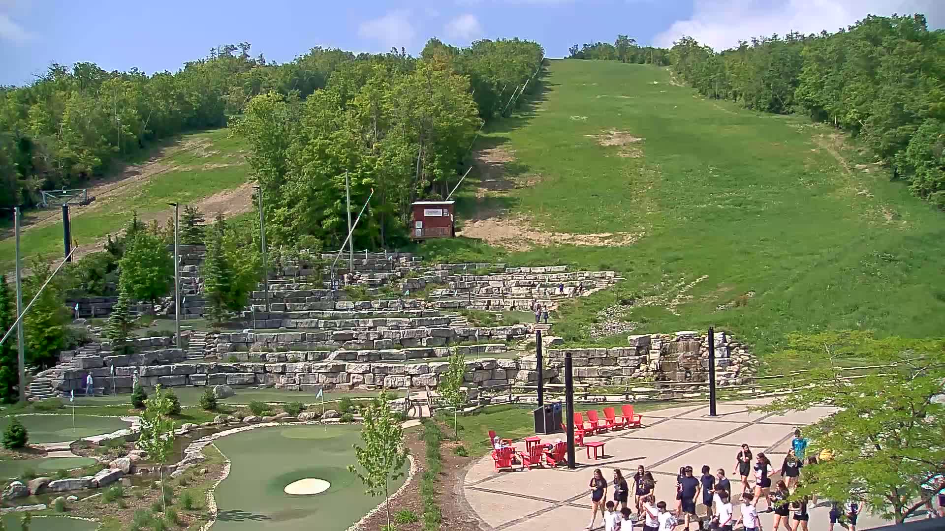 A grassy hillside with a ski lift and a stone mini-golf course below, under sunny skies.