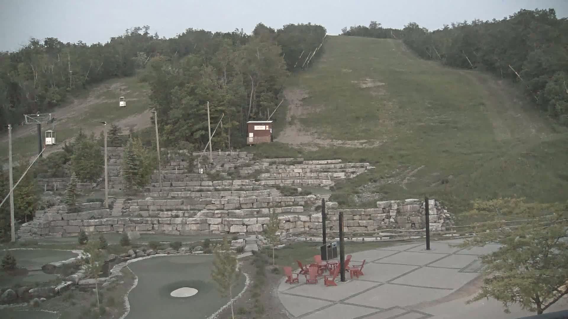 A grassy hillside with a ski lift and stone retaining walls, a paved area with red chairs and a table, and a putting green are visible under an overcast sky.