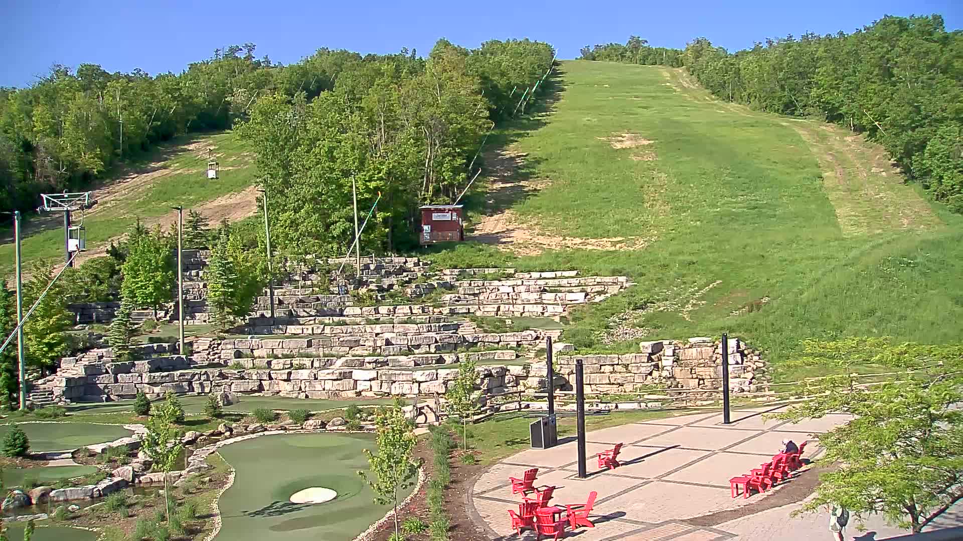 A grassy hillside with a chairlift and ski slope visible in the background, overlooks a paved area with red chairs and a mini golf course constructed from stone retaining walls on a sunny day.