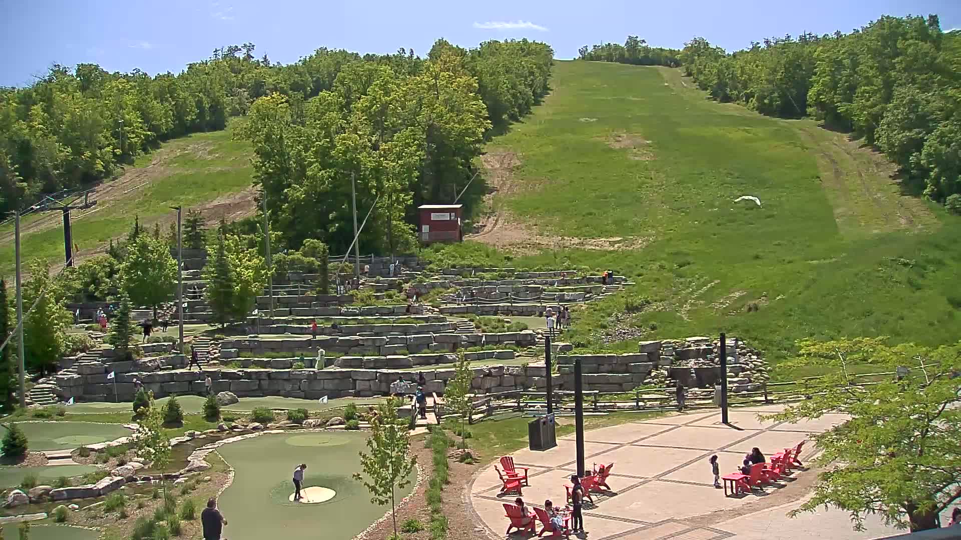 A grassy hillside slopes upward behind a stone-walled mini golf course, where people are playing, and a paved patio area with red chairs and people relaxing on a sunny day.