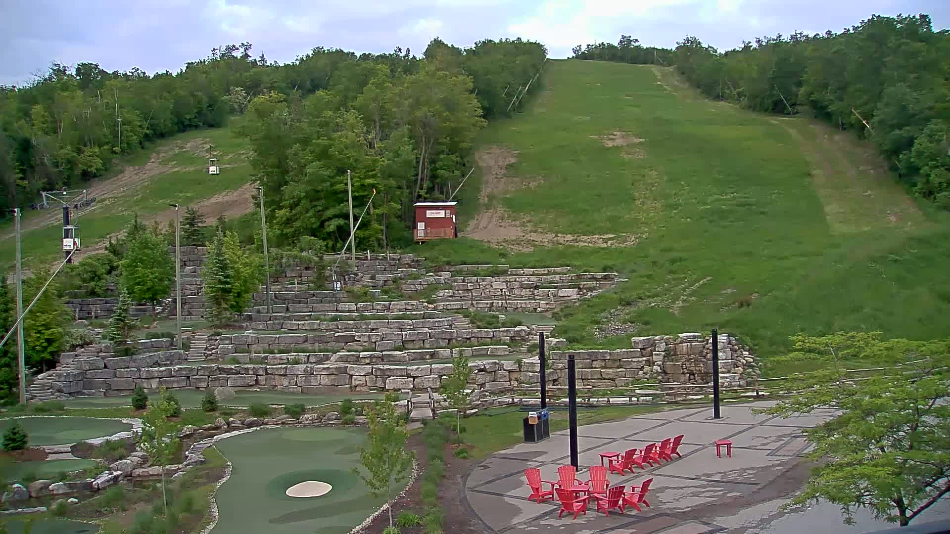 A grassy hillside with a ski lift, a stone amphitheater, a mini golf course, and red chairs on a patio under a cloudy sky.