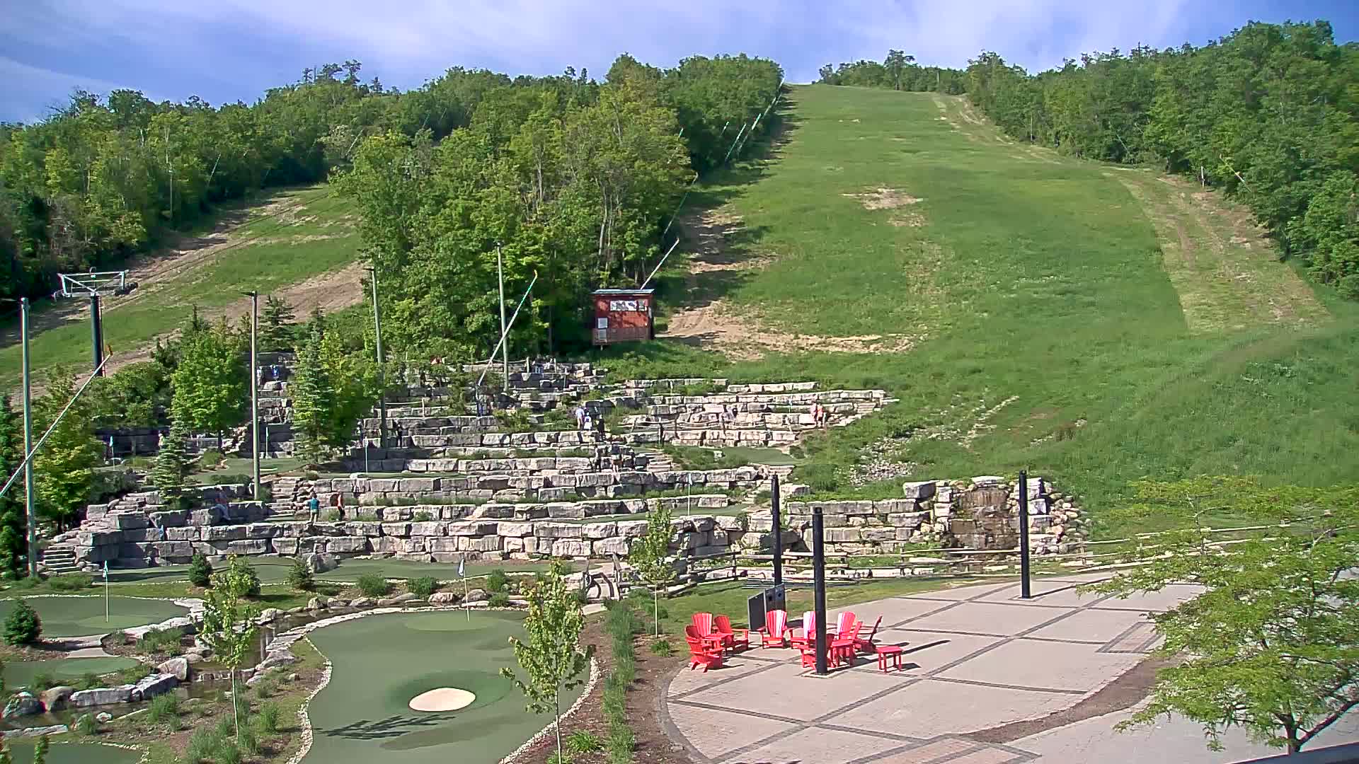 A grassy hillside with a ski lift, a stone amphitheater, a miniature golf course, and several red Adirondack chairs under a mostly sunny sky.