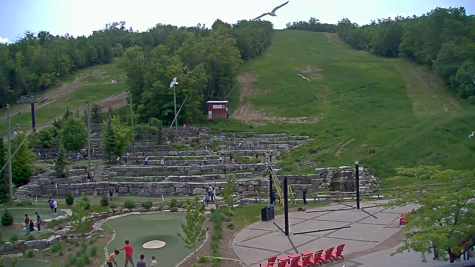 A grassy hillside with a ski lift, overlooks an outdoor mini golf course with people playing, under a partly cloudy sky.