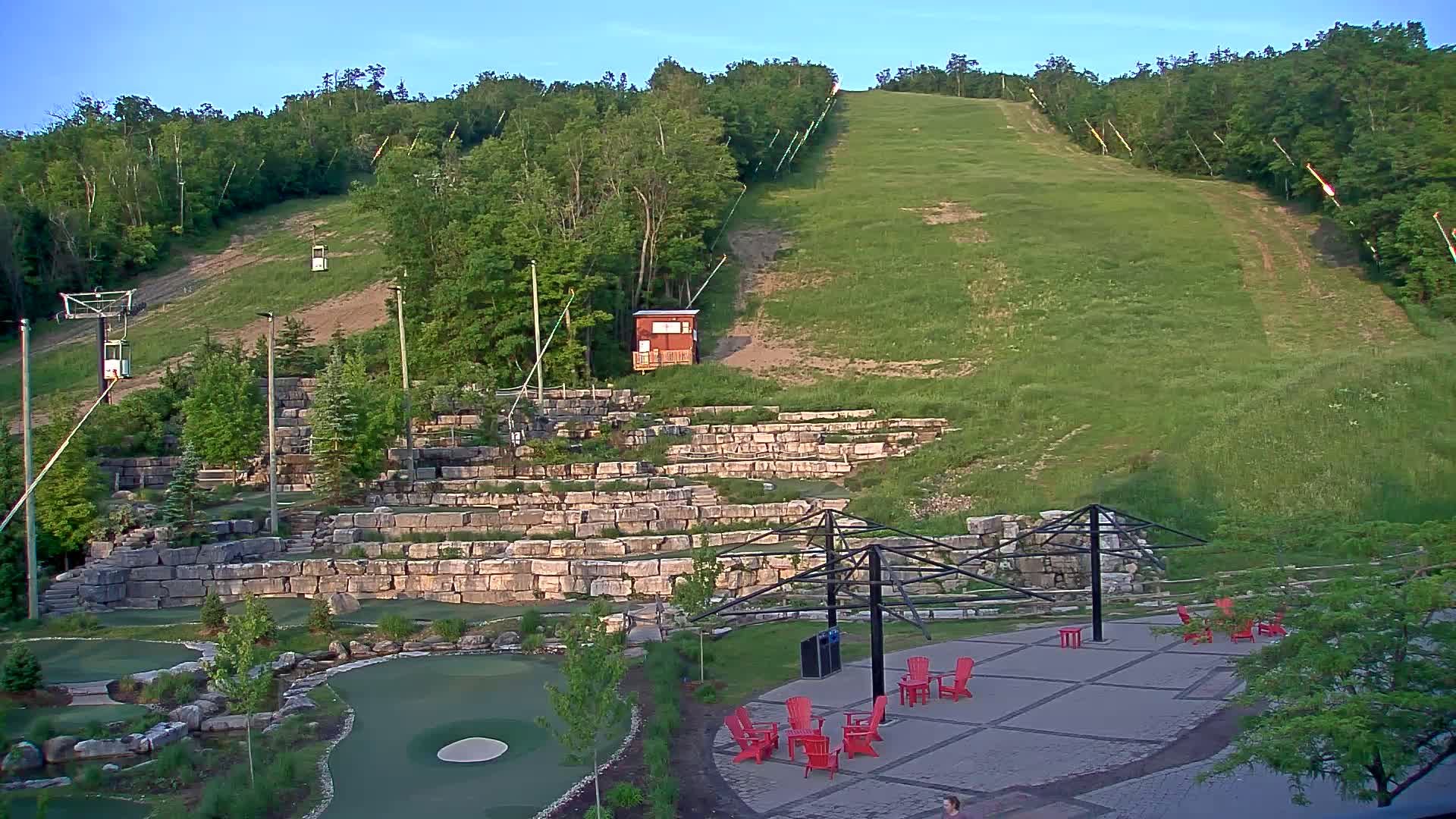 A grassy hill with a ski lift and a stone retaining wall features a mini-golf course and a patio area with red chairs beneath a clear, bright sky.