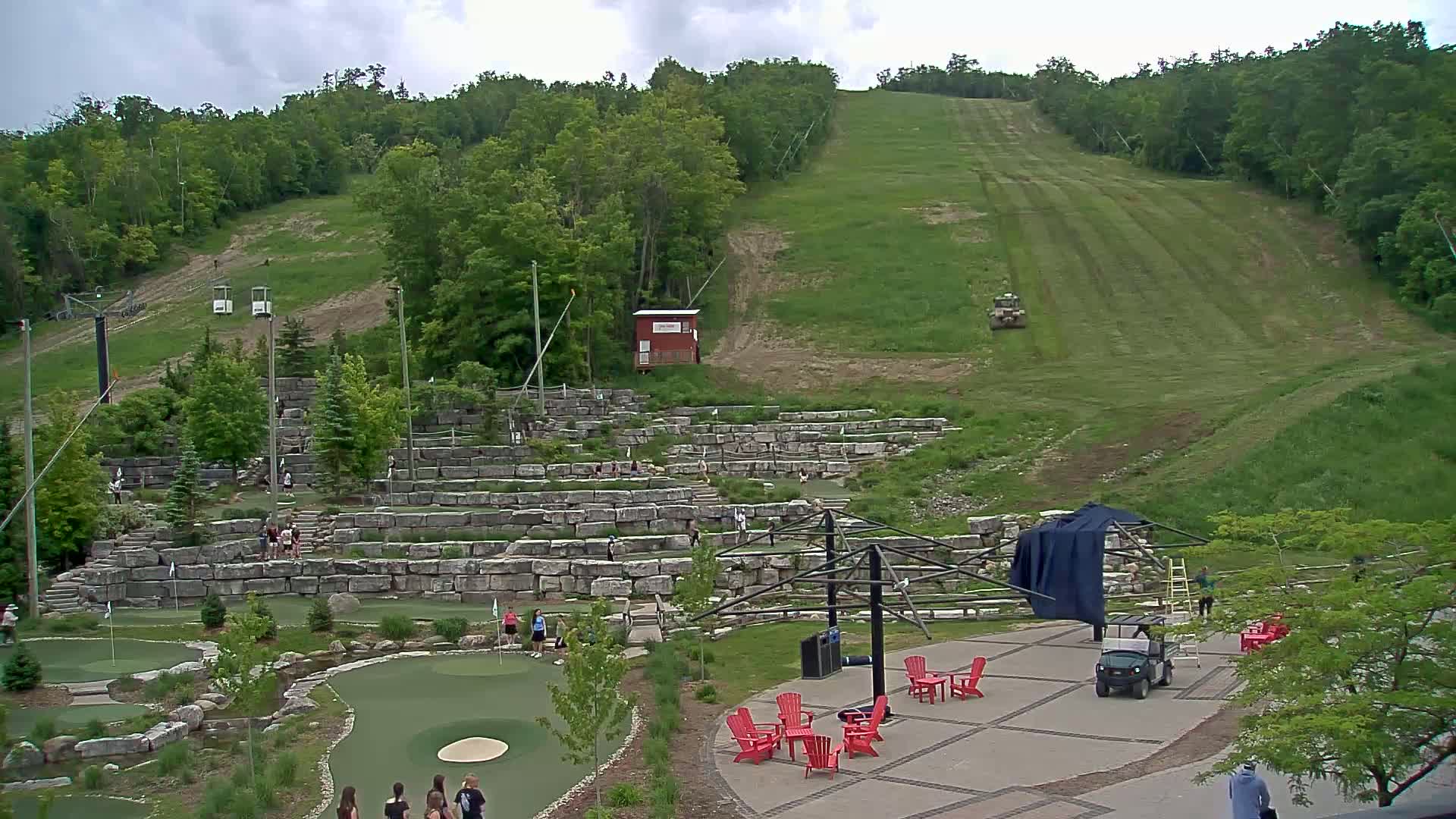 An outdoor scene under cloudy skies shows a grassy ski hill with a chairlift, a mini golf course built into stone terraces, and a paved area with red Adirondack chairs and a golf cart.