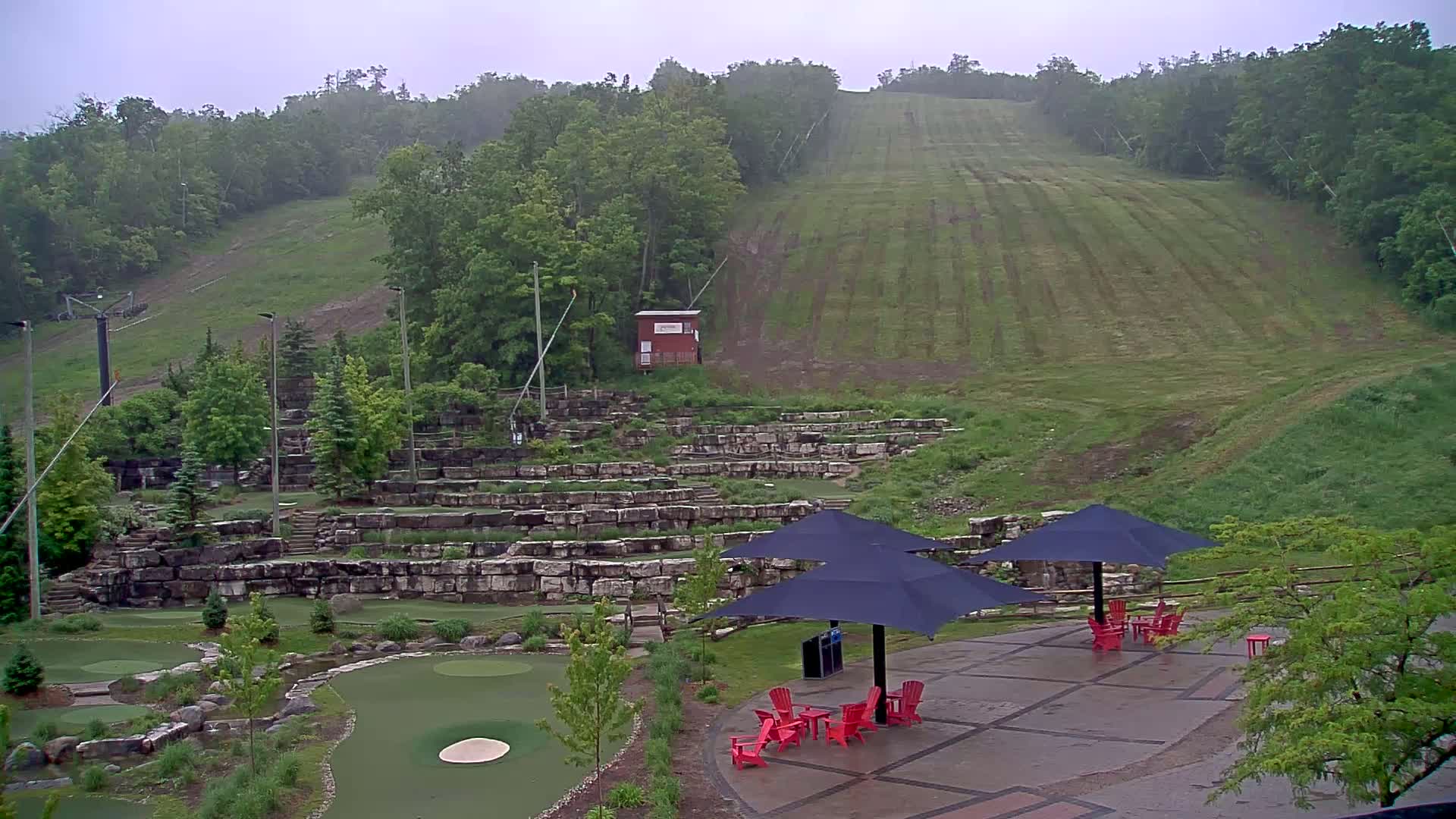 A rain-soaked outdoor mini-golf course with stone retaining walls sits below a grassy hill with a ski lift, under overcast skies.