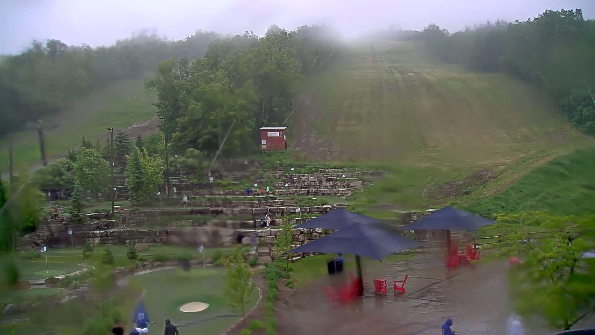A rain-soaked miniature golf course with a hill and trees in the background, under a misty, overcast sky.